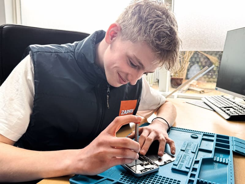 Jonge reparateur schroeft lachend een iPhone open op een blauwe ESD-mat in de werkplaats